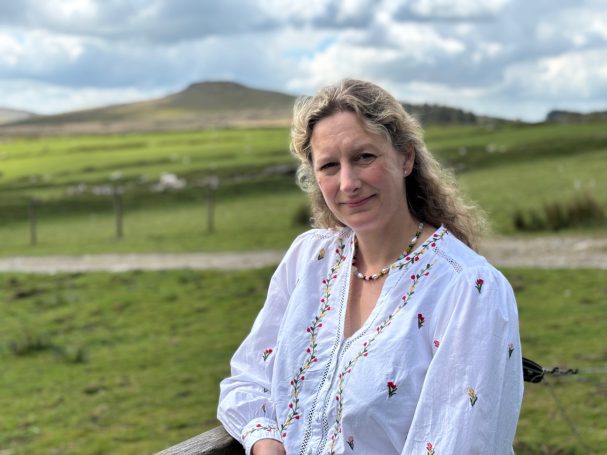 Woman in a white blouse with embroidery, set against a grassy landscape and hills.